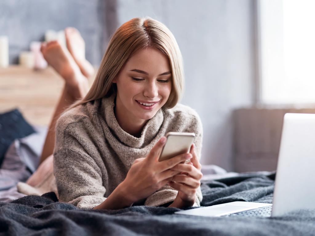 Delighted woman using smartphone and laptop in bed